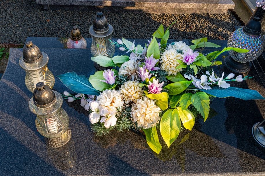 Grabstein mit Blumenarrangement in Weiß und Lila sowie mehreren Grablichtern aus Glas und Metall